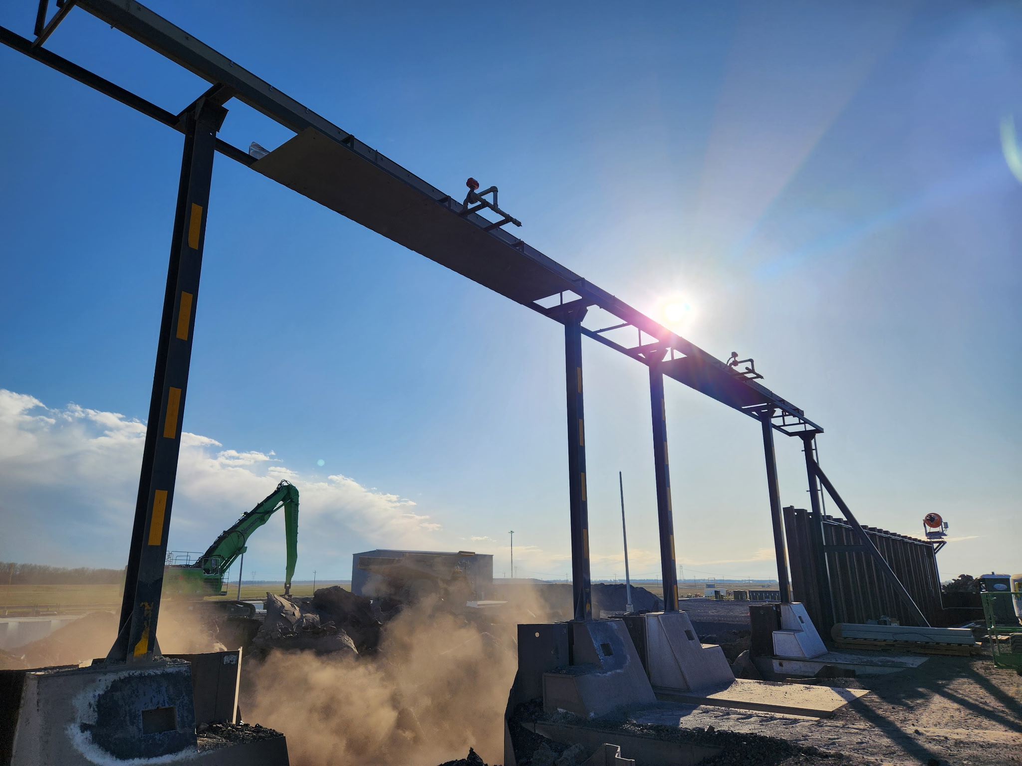 Overhead Pipe Truss & Heat Shield Installation Steel overhead pipe truss system installed over industrial dump station with reinforced concrete foundations and heavy equipment operating below.