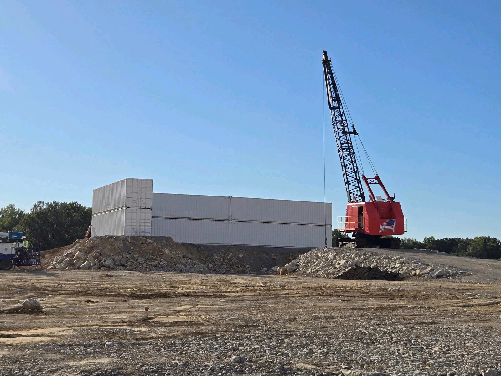 Stacked Shipping Container Protection Wall Installation Crane setting stacked 40-foot shipping containers for a welded protection wall at an industrial site, with containers positioned three high and three long on a compacted pad.